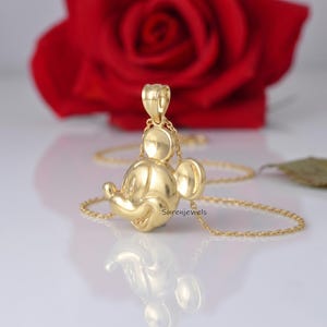 May include: A polished gold pendant shaped like the head of Mickey Mouse, suspended from a delicate gold chain. The pendant features a smooth, reflective surface with the iconic rounded ears and facial features of the character in a solid gold finish.