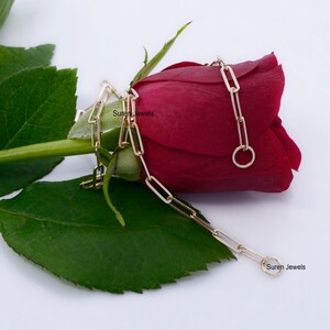 May include: A gold chain necklace with a rectangular link design. The chain is draped over a red rose bud. The text "Suren Jewels" appears twice in the image.