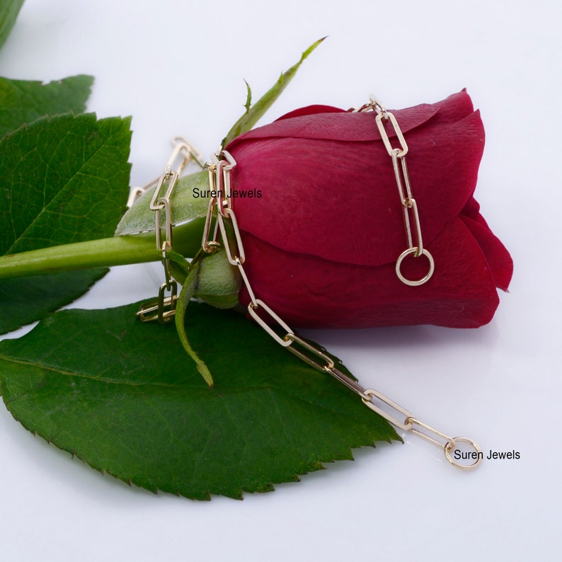 May include: A gold chain necklace with a rectangular link design. The chain is draped over a red rose bud. The text "Suren Jewels" appears twice in the image.