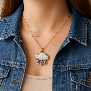 Pode incluir: Um colar de corrente dourada com um pingente em forma de nuvem com um centro branco iridescente e três gotas azuis pendentes. O pingente está suspenso por uma delicada corrente de metal dourado, com o texto Surenjewels visível na imagem.