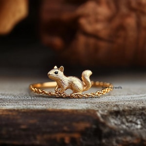 May include: A gold ring with a detailed squirrel charm. The squirrel is facing to the right and has a black eye. The ring is made of a delicate chain.