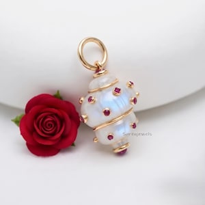 Könnte beinhalten: Ein goldener Anhänger in Form einer Muschel mit einer glatten, weiß schimmernden Oberfläche, verziert mit kleinen, runden, leuchtend roten Edelsteinen in Goldfassungen, neben einer dunkelroten Rose mit grünen Blättern.