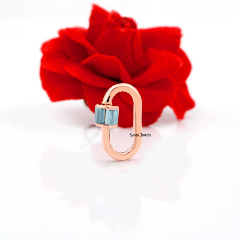 May include: Rose gold charm featuring a light blue gemstone. The charm is shaped like a sideways oval with a small loop at the top. The charm is displayed against a white background.
