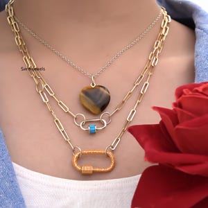 May include: A layered gold chain necklace featuring a tiger's eye heart pendant and a turquoise blue charm. The necklace is worn by a person with a red rose in the background.