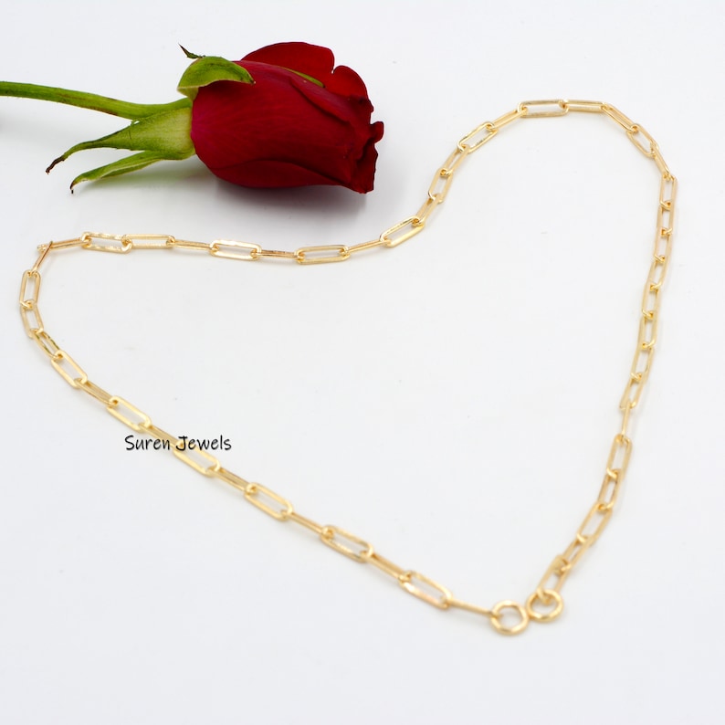 May include: A gold chain necklace with a delicate, rectangular link design. The chain is displayed on a white background with a red rose.