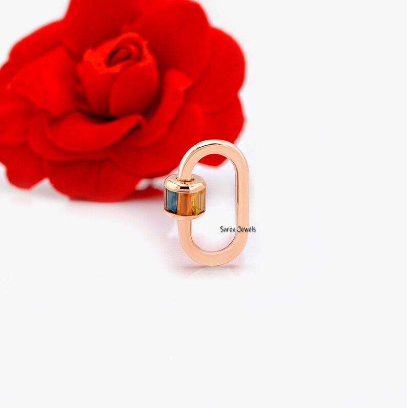 May include: A rose gold oval-shaped charm with a multi-coloured gemstone set in the centre. The charm is a lobster clasp style and says "Suren Jewels" on the side.
