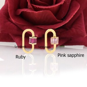 May include: Two gold charm earrings with a rectangular shaped stone. The earring on the left has a red ruby stone and the earring on the right has a pink sapphire stone. The earrings are on a red rose background.