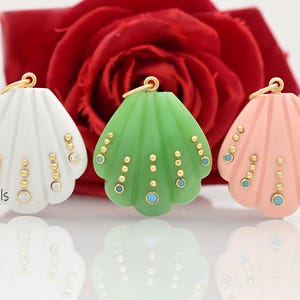May include: Three shell-shaped pendants in white, green, and peach, each featuring a gold-tone metal loop at the top. The surface of each pendant is decorated with vertical rows of small gold-tone beads and embedded circular stones.