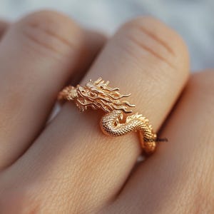 Op de afbeelding: Een gouden drakenring met een gedetailleerd ontwerp. De kop van de draak is naar voren gericht, met de bek open en de vleugels uitgespreid. De ring wordt aan een vinger gedragen.