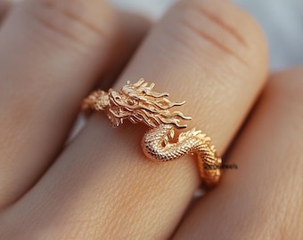May include: A gold-toned dragon ring with a detailed design. The dragon's head is facing forward with its mouth open and its wings spread out. The ring is worn on a person's finger.
