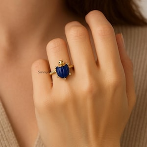 Può includere: Un anello in oro con una tartaruga scolpita in lapislazzuli blu come elemento centrale, dotata di una piccola testa dorata e quattro minuscole pietre bianche d'accento sulle zampe. La fascia presenta una trama sottile e il testo Surenjewels è visibile vicino alla tartaruga.