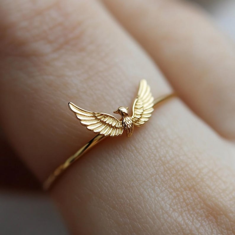 May include: A gold ring with a bird design. The bird has its wings spread out and is facing forward. The ring is on a person's finger.