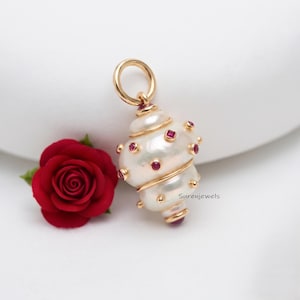 May include: A gold pendant featuring a white baroque pearl shaped like a shell, accented with small square-cut red rubies set in gold. The pendant includes a circular gold bail at the top and is positioned next to a deep red rose flower.