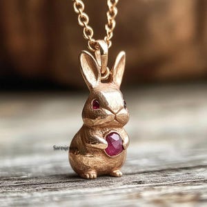 Może przedstawiać: Złoty naszyjnik z zawieszką w kształcie królika z czerwonym kamieniem szlachetnym. Królik trzyma kamień szlachetny w łapkach.