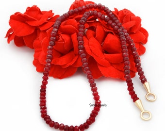 May include: A red gemstone necklace with gold clasps. The necklace is made of small, faceted beads and is displayed on a bed of red roses.