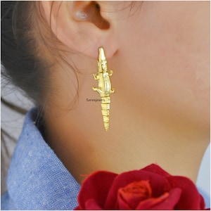 May include: Gold alligator earrings. The alligator is detailed with textured scales and has a long tail.