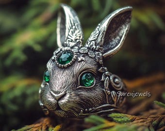 May include: A silver rabbit ring with green gemstone eyes and intricate floral details. The ring is set against a green background.