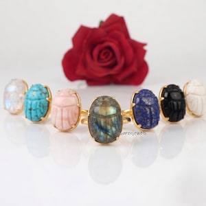 May include: A collection of seven gold-toned rings featuring oval-shaped scarab-carved gemstones in various colors including iridescent white, turquoise, pale pink, labradorite, deep blue lapis lazuli, solid black, and opaque white stone.
