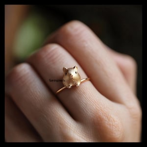 Puede incluir: Anillo dorado con un diseño detallado de cobaya en tres dimensiones. El anillo tiene una banda fina y delicada, y la cobaya es el punto focal, con pelaje y rasgos faciales visibles.