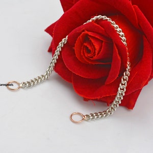 Puede incluir: Un brazalete de cadena de plata con dos pequeños anillos de oro rosa. El brazalete está sobre una rosa roja.