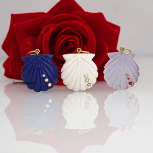 May include: Three shell-shaped pendants featuring a gold jump ring at the top. The left pendant is deep blue with three clear stones, the centre is white with three neutral stones, and the right is pale lavender with three small red stones.