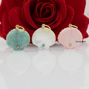 May include: Three seashell pendants in shades of green, white, and pink, each with a gold-colored loop and a small gemstone accent. The pendants are set against a blurred background of a red rose.
