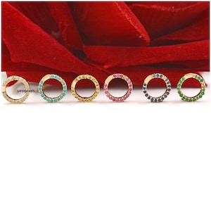 Puede incluir: Seis pendientes de aro de oro con un círculo de piedras preciosas de colores. Los pendientes están dispuestos en fila de izquierda a derecha: blanco, turquesa, amarillo, rosa, azul y verde.