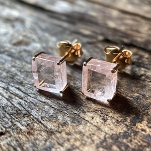 May include: A pair of square-cut, pale pink gemstone stud earrings with gold-coloured settings. The earrings are presented on a weathered wooden surface, highlighting their delicate colour and design. The earrings are a classic jewellery item.