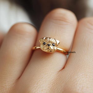 Puede incluir: Anillo de oro con un diseño detallado de cabeza de carlino. El anillo se lleva en el dedo de una persona.