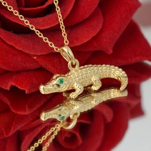 Op de afbeelding: Een goudkleurige ketting met een gedetailleerde alligatorhanger met groene edelsteenogen. De hanger hangt aan een fijne ketting. De afbeelding staat tegen een rode achtergrond.
