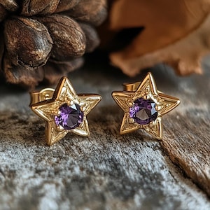 Puede incluir: Un par de pendientes de botón con forma de estrella en tono dorado, cada uno con una piedra preciosa redonda de color púrpura en el centro. La superficie metálica de las estrellas tiene un acabado texturizado y grabado con cierres de presión.