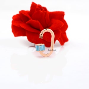 May include: Rose gold carabiner charm with a light blue baguette-cut gemstone. The charm is on a white background with a red rose in the background. "Siren Jewels" is printed on the charm.