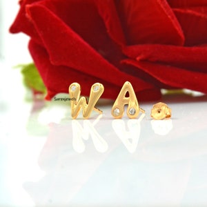 Może przedstawiać: Złote kolczyki sztyfty z literami "WA" z małymi, przezroczystymi kamieniami szlachetnymi. Kolczyki są prezentowane na odblaskowej powierzchni z rozmytą czerwoną różą w tle. Kolczyki mają sztyft i zapięcie.