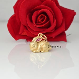 Könnte beinhalten: Ein goldfarbener Kaninchenanhänger mit strukturierter Oberfläche und kleinen rosa Steinen in den Ohren und auf dem Rücken. Der Anhänger verfügt über einen runden Ring an der Oberseite. Der Text Surenjewels ist rechts neben dem Kaninchen sichtbar.
