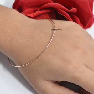 May include: A thin, rose gold bangle bracelet on a person's wrist. The bracelet is simple and elegant, with a smooth, polished finish.