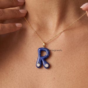 May include: A gold chain necklace with a blue letter "R" pendant. The pendant is a deep blue color with two small, round, clear gemstones. The necklace is worn around the neck.