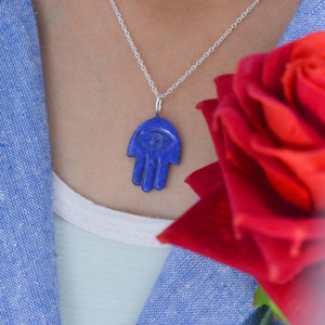 Puede incluir: Un collar de cadena de plata con un colgante de mano de hamsa azul. La mano de hamsa tiene un ojo azul en el centro. El collar lo lleva una persona con una rosa roja en el fondo.