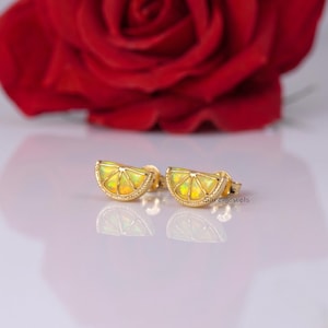 Può includere: Un paio di orecchini a lobo in oro a forma di spicchi di agrumi, con inserti in opale giallo e un design segmentato. Gli orecchini presentano un bordo in oro lucido e sono posizionati in modo da mostrare il loro riflesso su una superficie.