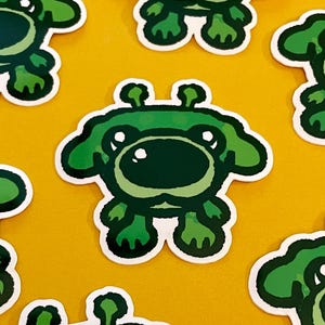May include: Seven green cartoon frog stickers with white outlines are arranged on a yellow background. Each sticker features a frog with a large, dark green nose, big eyes, and a small crown. The stickers are a fun, whimsical design.