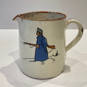 Może przedstawiać: Biały dzbanek ceramiczny z brązową krawędzią i ręcznie malowanym wzorem przedstawiającym żołnierza w niebieskim płaszczu trzymającego karabin. Żołnierz stoi w śnieżnym krajobrazie.