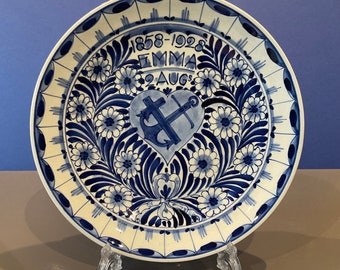 May include: A white ceramic plate with a blue and white floral design. The center of the plate features a heart with an anchor and cross inside. The text "1858-1928 EMMA 9 AUG" is written above the heart.