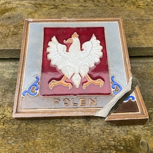 Puede incluir: Un azulejo de cerámica roto con un águila blanca y la palabra "POLEN" en letras doradas. El águila está sobre un fondo rojo con detalles dorados. El azulejo está rodeado por un borde azul y blanco.