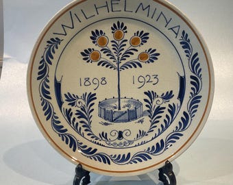 May include: A white ceramic plate with a blue and gold design. The plate features a tree with fruit, a fence, and the text "WILHELMINA 1898 1923".