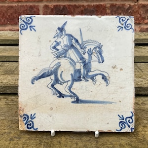 Puede incluir: Un azulejo de Delft azul y blanco que representa a un hombre a caballo. El hombre lleva un sombrero y sostiene una espada. El caballo se levanta sobre sus patas traseras.