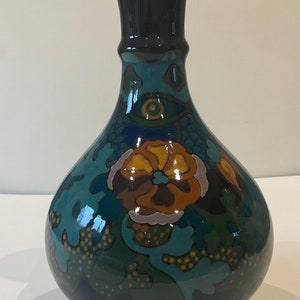 Puede incluir: Un jarrón de cerámica azul oscuro con una base ancha y un cuello estrecho. El jarrón está decorado con un patrón floral en tonos de verde, marrón, amarillo y azul.