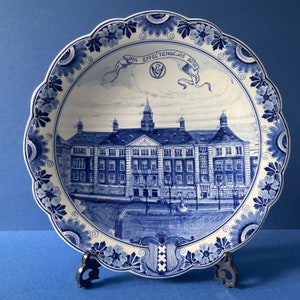 Peut inclure: Une assiette en faïence de Delft bleue et blanche représentant un bâtiment et le texte "1914 Effectenbeurs 1919". L'assiette est ornée d'une bordure florale et d'un motif central représentant un bâtiment avec une tour d'horloge.