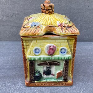 Op de afbeelding: Een keramische huisvormige container met een rieten dak en schoorsteen. De gele muren hebben een haardontwerp met de tekst "YE OLDE FIRESIDE" aan de basis. Het dak is versierd met bloemmotieven.