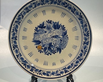 May include: A white ceramic plate with a blue and white floral design. The plate has a clock face with Roman numerals and a banner that reads "Prinses Irene 5 Aug 1959".