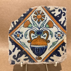 May include: Fragment of a decorative tile featuring a central vase with flowers and foliage. The tile has a white background with orange, blue, and green floral designs. The tile is framed by a blue and orange border.