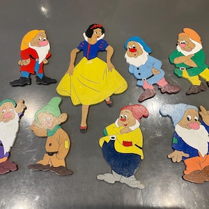 以下が含まれることがあります： 青と黄色のドレスを着た白雪姫と、7人の小人が描かれた8つの手描き木製カットアウトのセット。各フィギュアは、赤、緑、紫、オレンジなどの鮮やかな色で彩られ、表情豊かに仕上げられています。